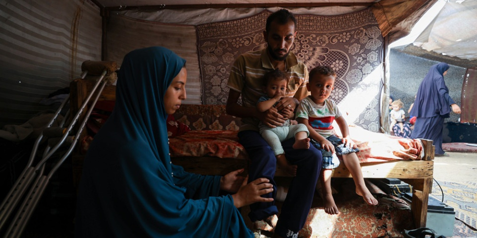 無國界醫生目睹加薩當地生活條件侵害人民基本尊嚴。巴索爾一家因戰火逃離拉法（Rafah），目前住在臨時帳篷，他抱著兩個孩子，妻子正在為他按摩截肢的腿，以緩解幻肢痛。數月以前，他在嘗試領取物資時中彈，失去了一條腿。照片攝於2025年10月9日。